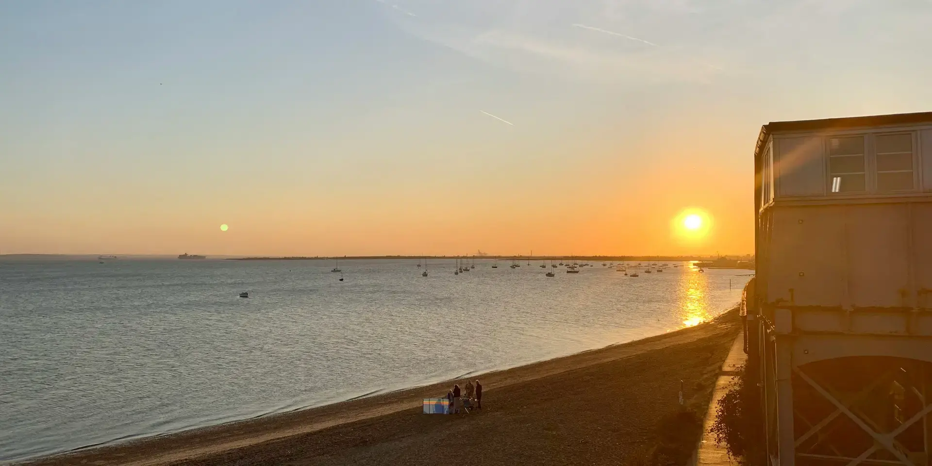 Golden sunset over a calm sea, with small boats scattered on the horizon and silhouettes of people on a sandy beach