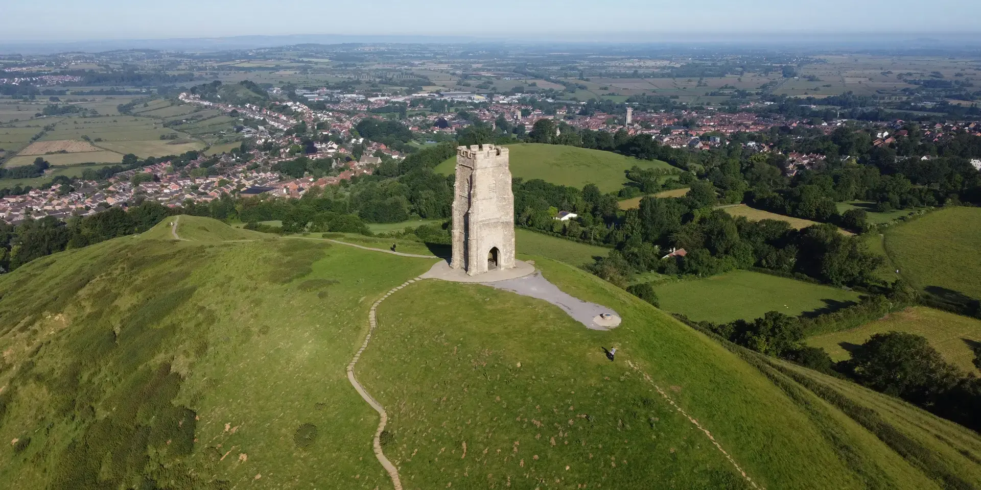 Aerial view of Glastonbury Tor with a stone tower on a green hill, overlooking a village and countryside