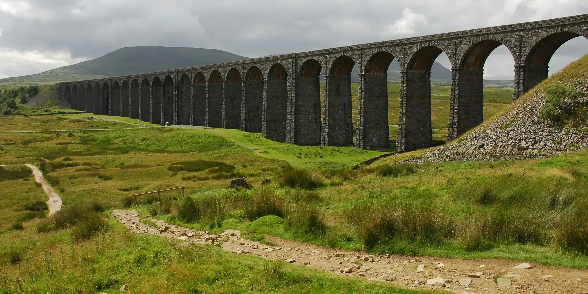 Stone viaduct with arches spans green moorland under cloudy skies, a dirt path winds towards the iconic structure