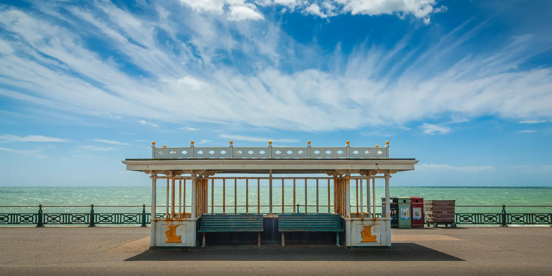 Seaside promenade with a vintage shelter, turquoise sea, and a bright blue sky with wispy clouds