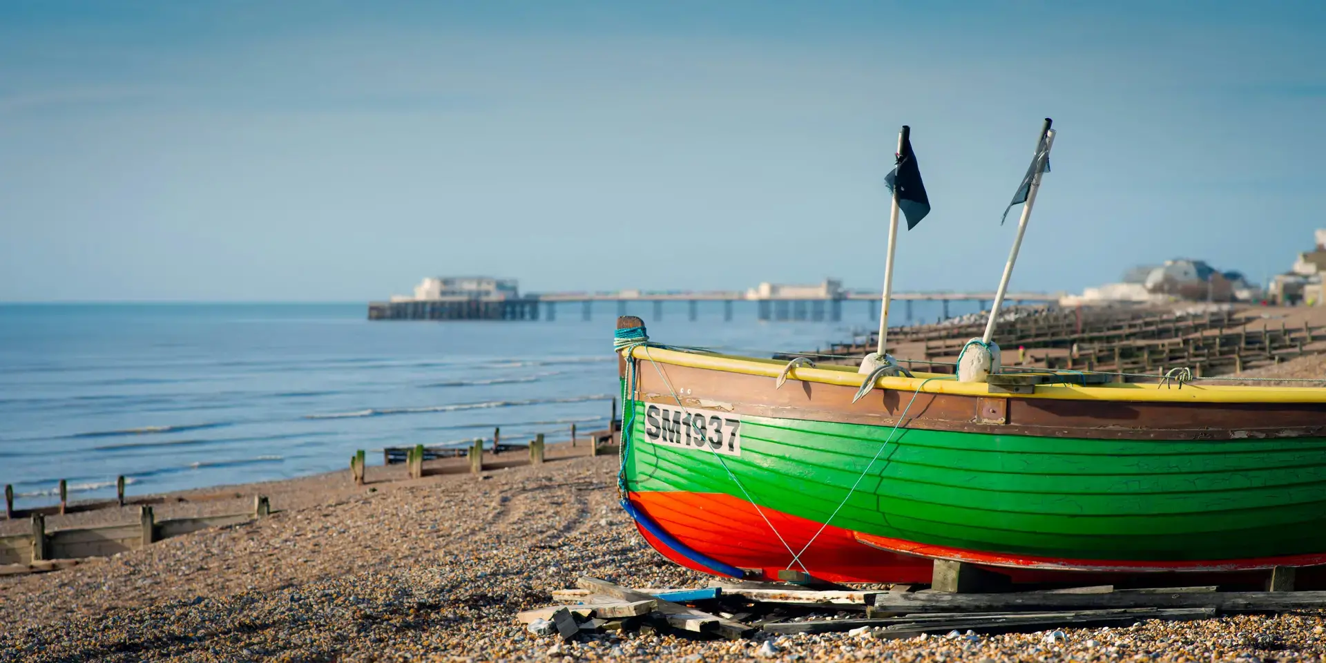 Vibrant fishing boat rests on a pebbled beach with a calm sea and distant pier, capturing a serene coastal morning scene