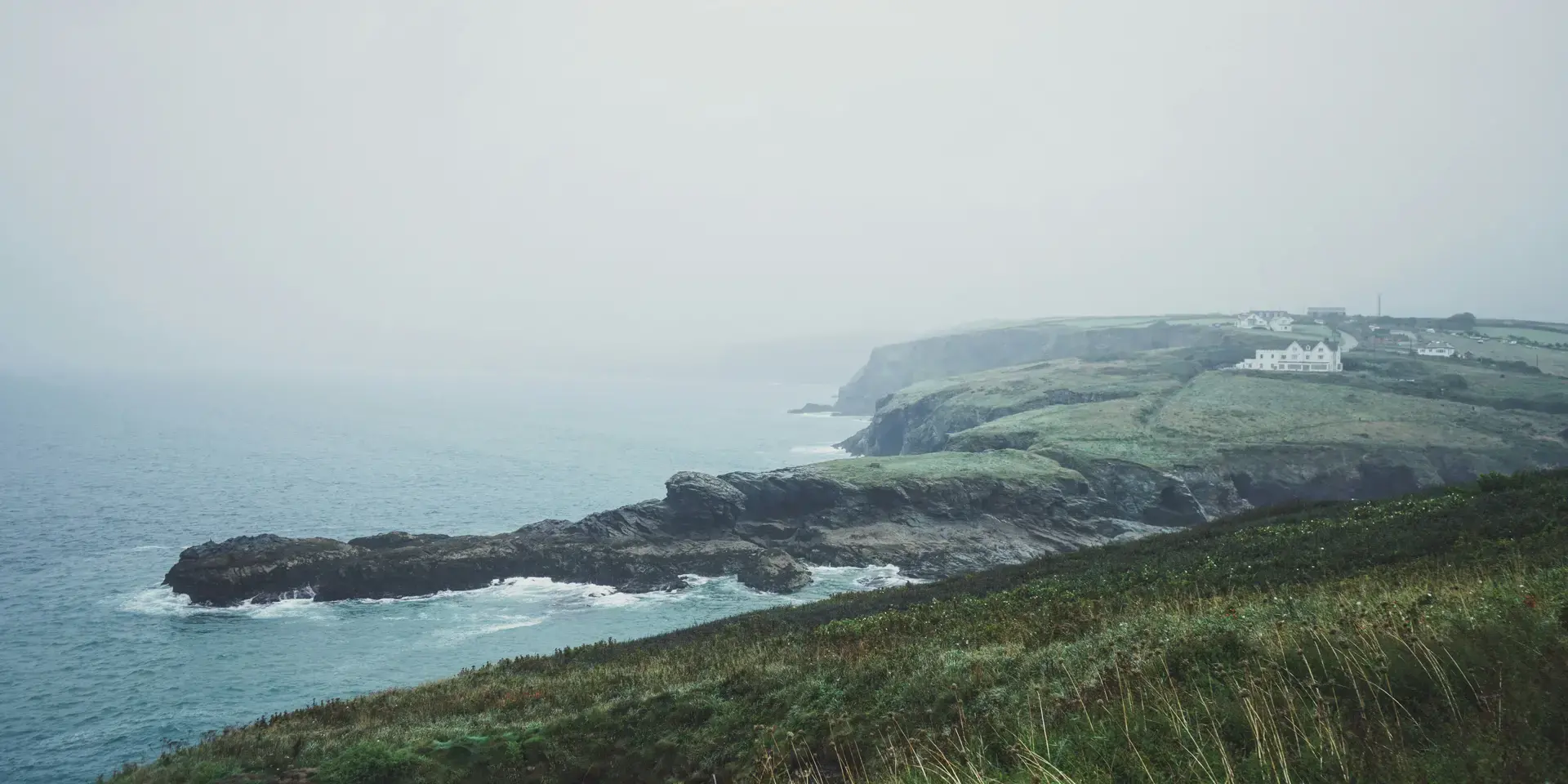 Misty coastal view with rugged cliffs and green fields, distant white cottages adding charm to the serene, foggy shoreline