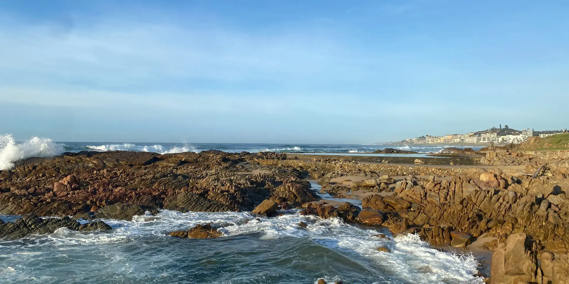 Rocky coastline with waves crashing against the shore, distant cityscape under a clear blue sky