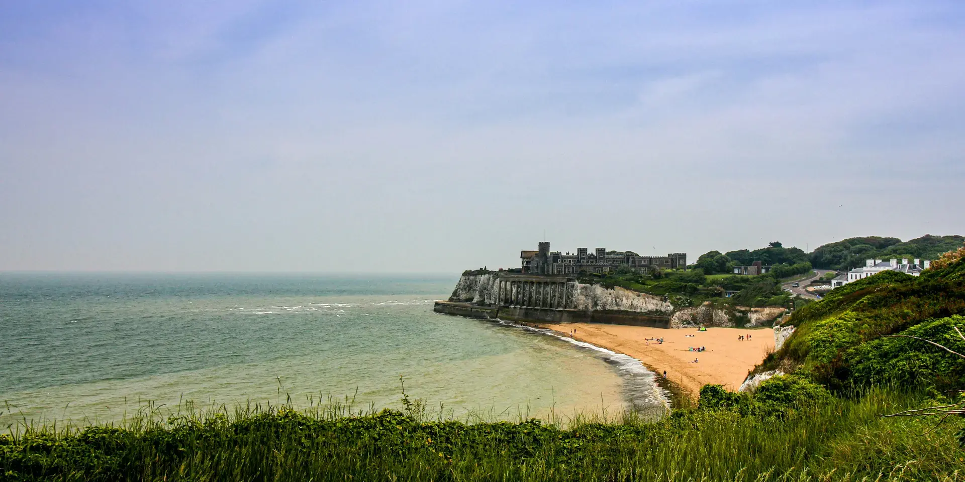 Coastal view with sandy beach, cliffs, and historic building overlooking calm sea under a hazy sky