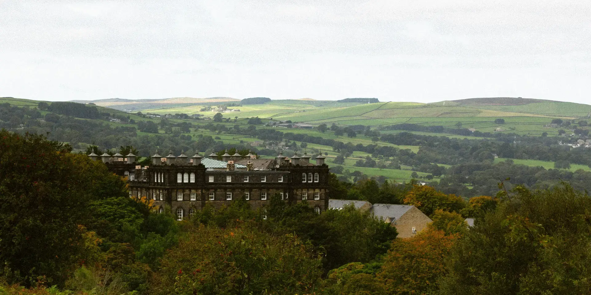Historic stone building nestled in lush greenery, overlooking the rolling countryside of Yorkshire under a cloudy sky