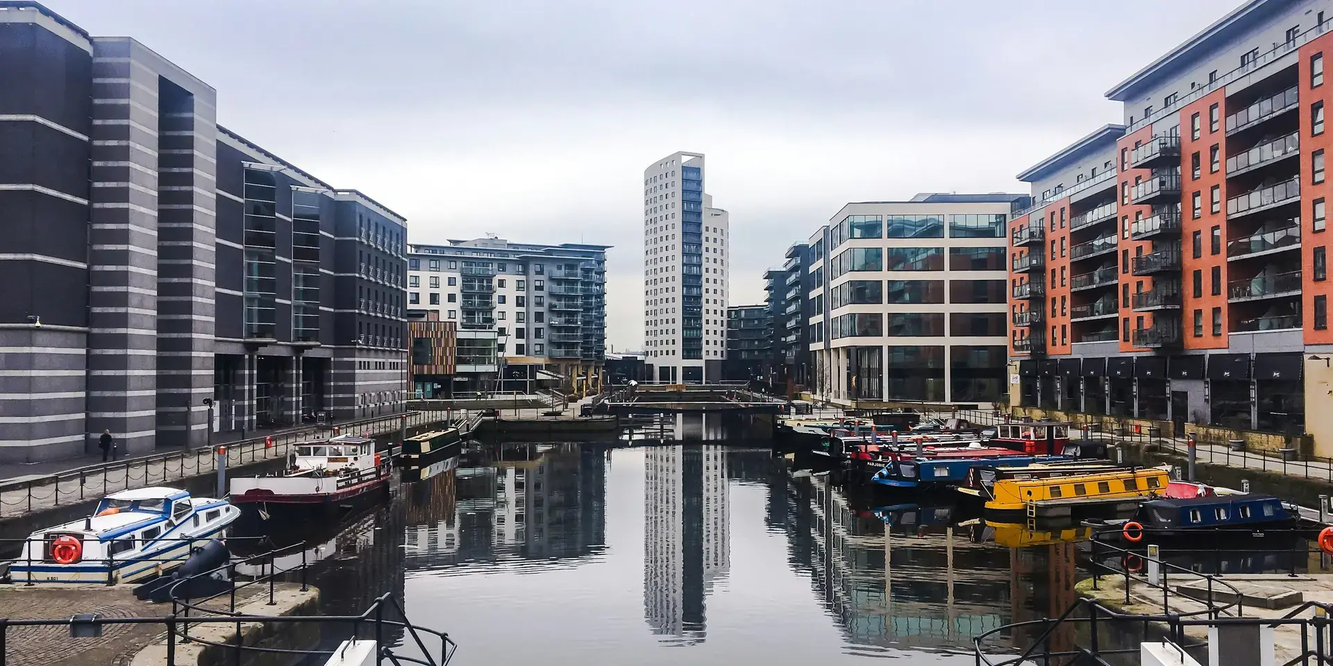 Canal boats docked along a calm waterway, framed by modern buildings under a cloudy sky. A peaceful urban scene