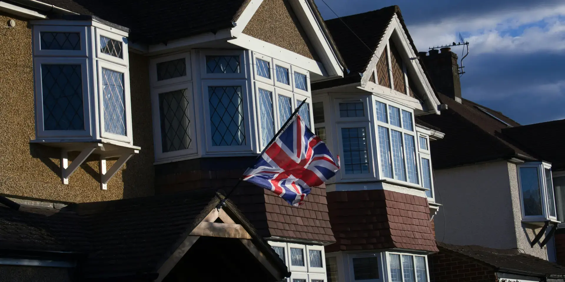 A Union Jack flag flutters in front of a row of classic British suburban houses under a dramatic evening sky