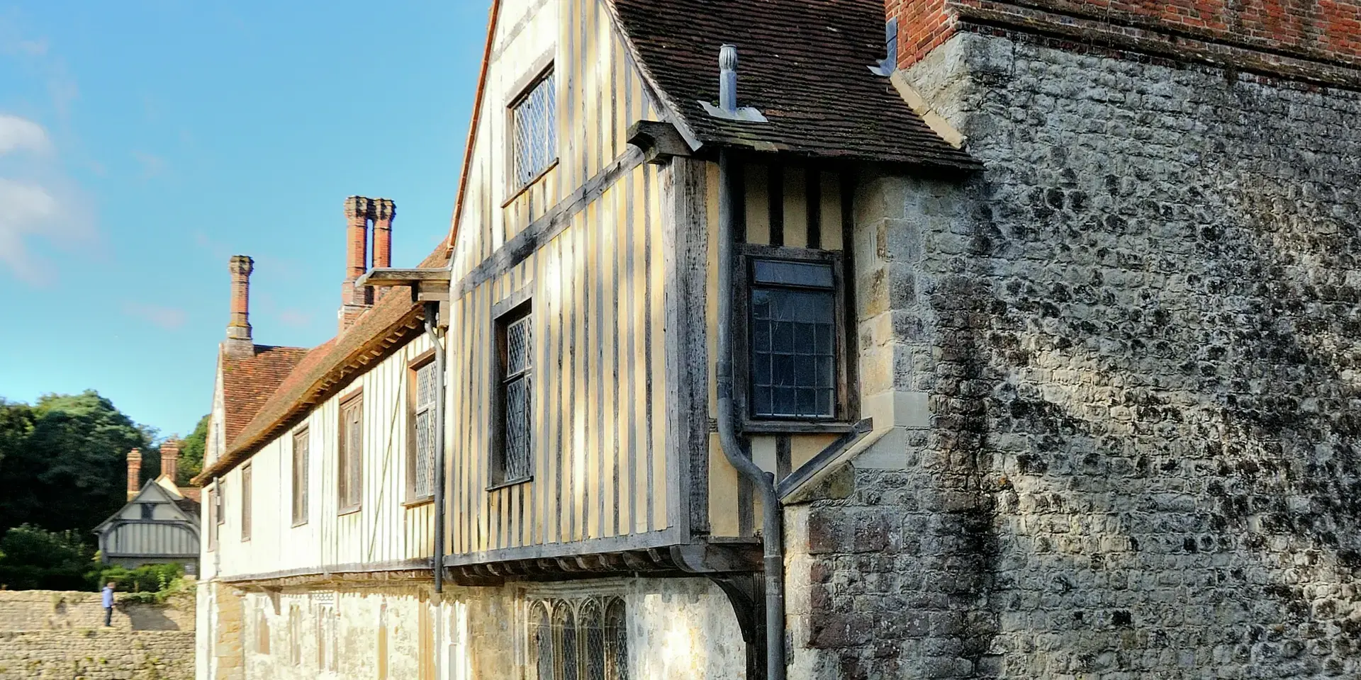 Tudor-style building with timber framing and tall brick chimneys reflecting in a calm moat under a blue sky