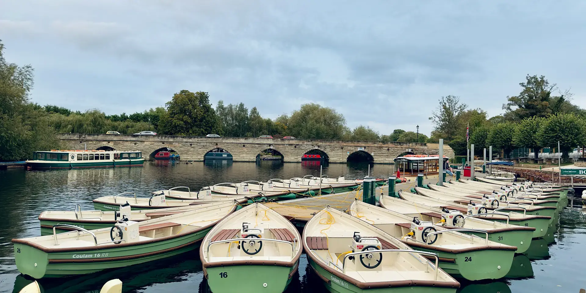 Cream and green boats docked near a stone bridge, reflecting on calm waters under a cloudy sky with trees in the background