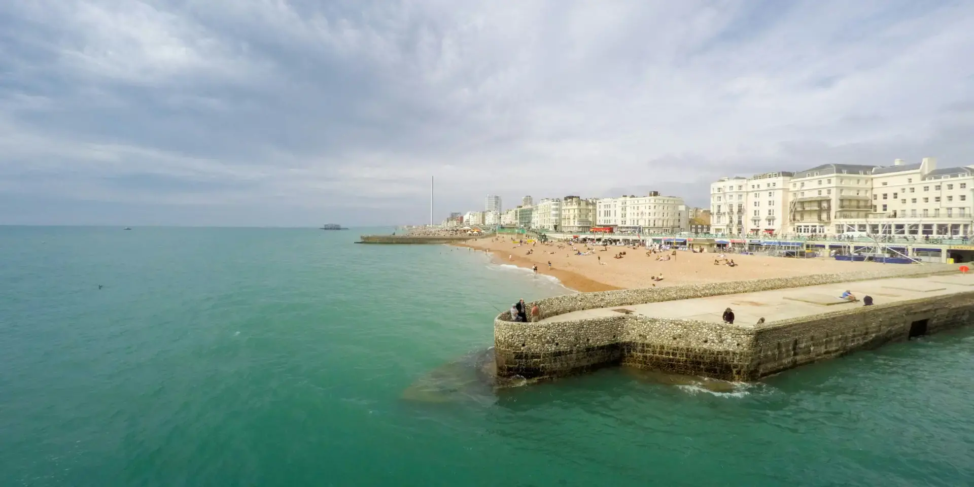 Coastal view with turquoise water, a stone pier, and a sandy beach lined with classic white buildings under a cloudy sky