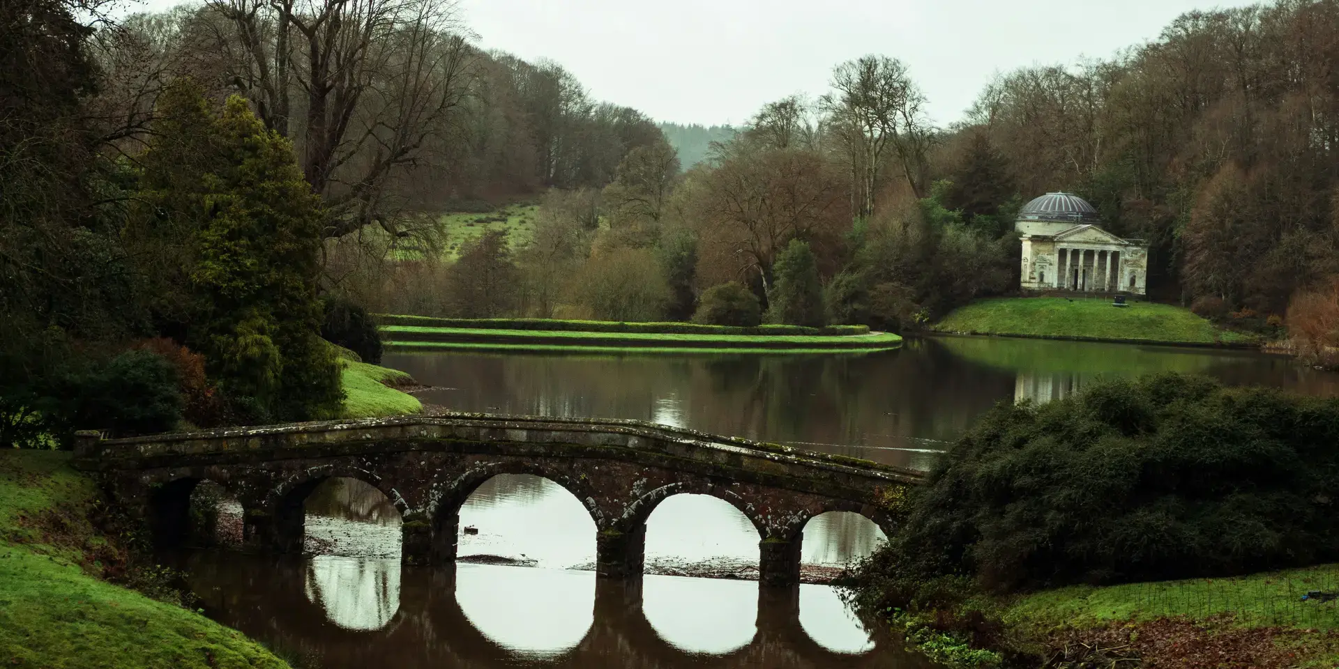 Stone bridge over a serene lake with a domed temple in the background, surrounded by lush greenery and peaceful scenery