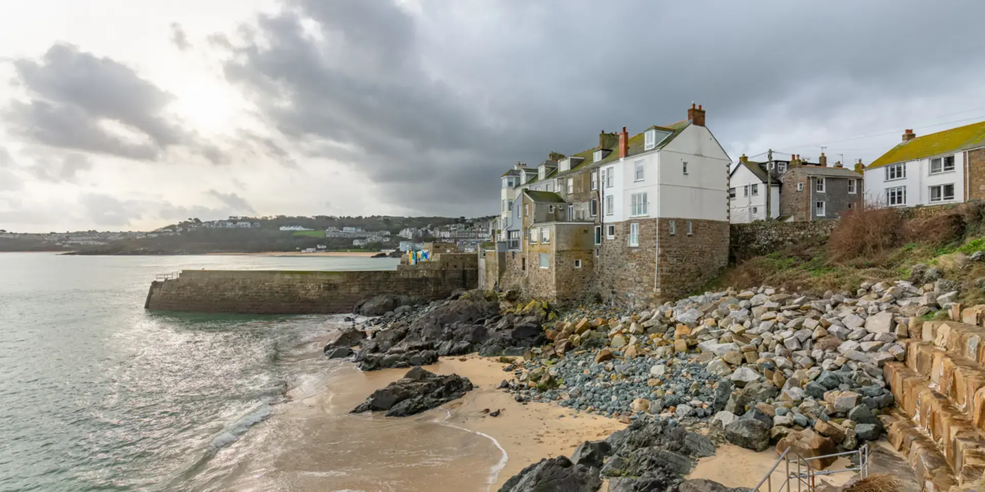 Stone houses on a coastal cliff overlooking a sandy beach and calm sea under a cloudy sky