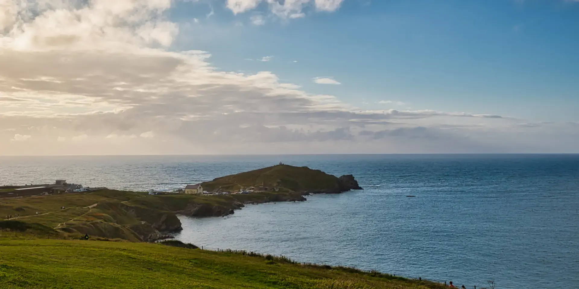 Grassy coastal headland extending into the sea under a partly cloudy sky with distant ocean views