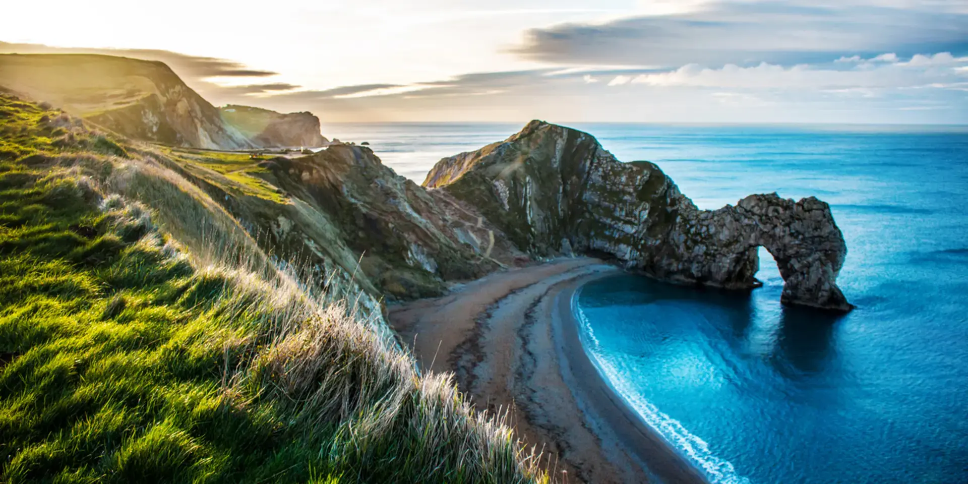 Scenic coastal view of a natural rock arch over blue water, with grassy cliffs under a bright sky.