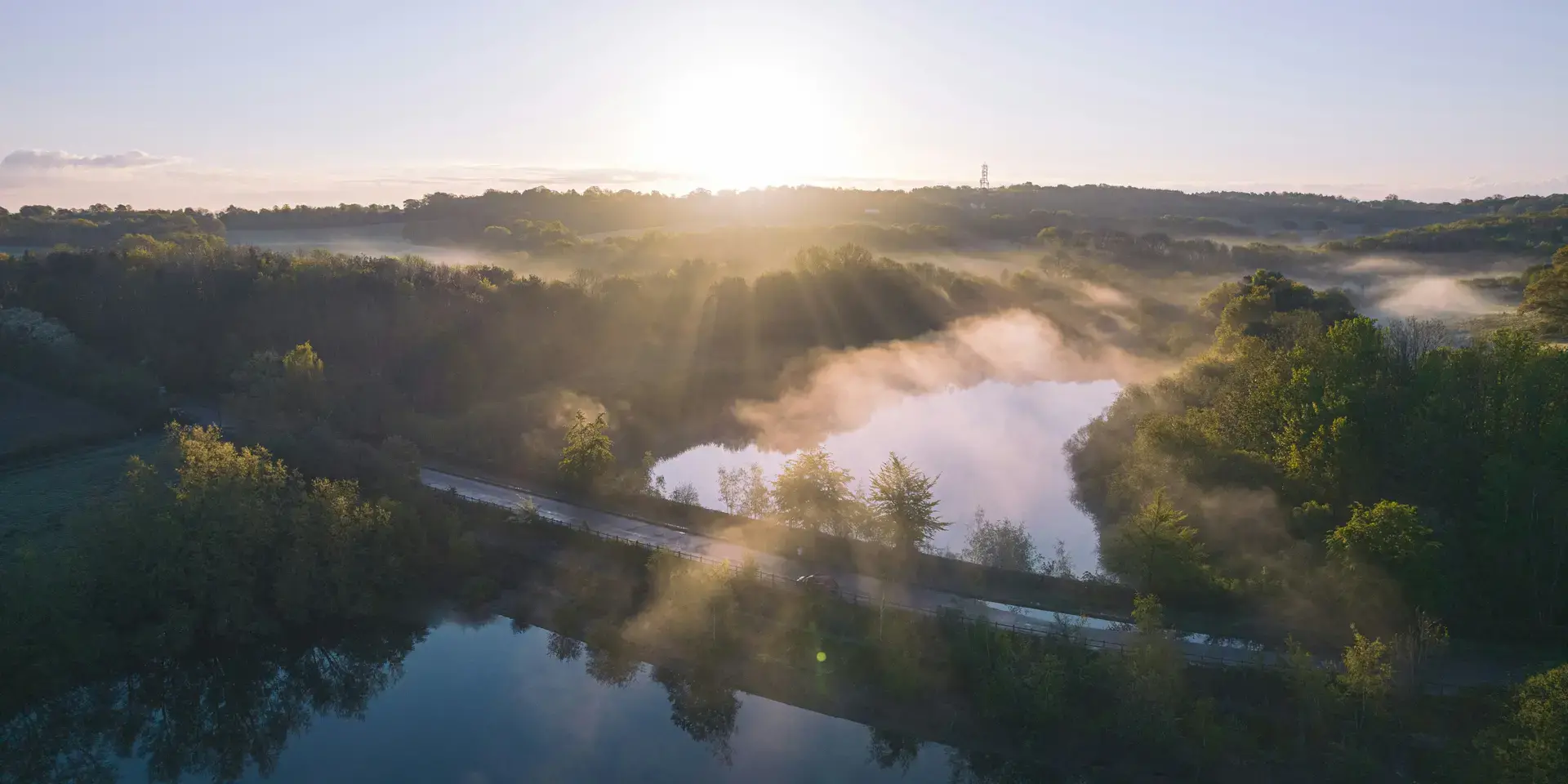 Serene sunrise over a misty forest with a reflective lake and a winding road cutting through the lush greenery.