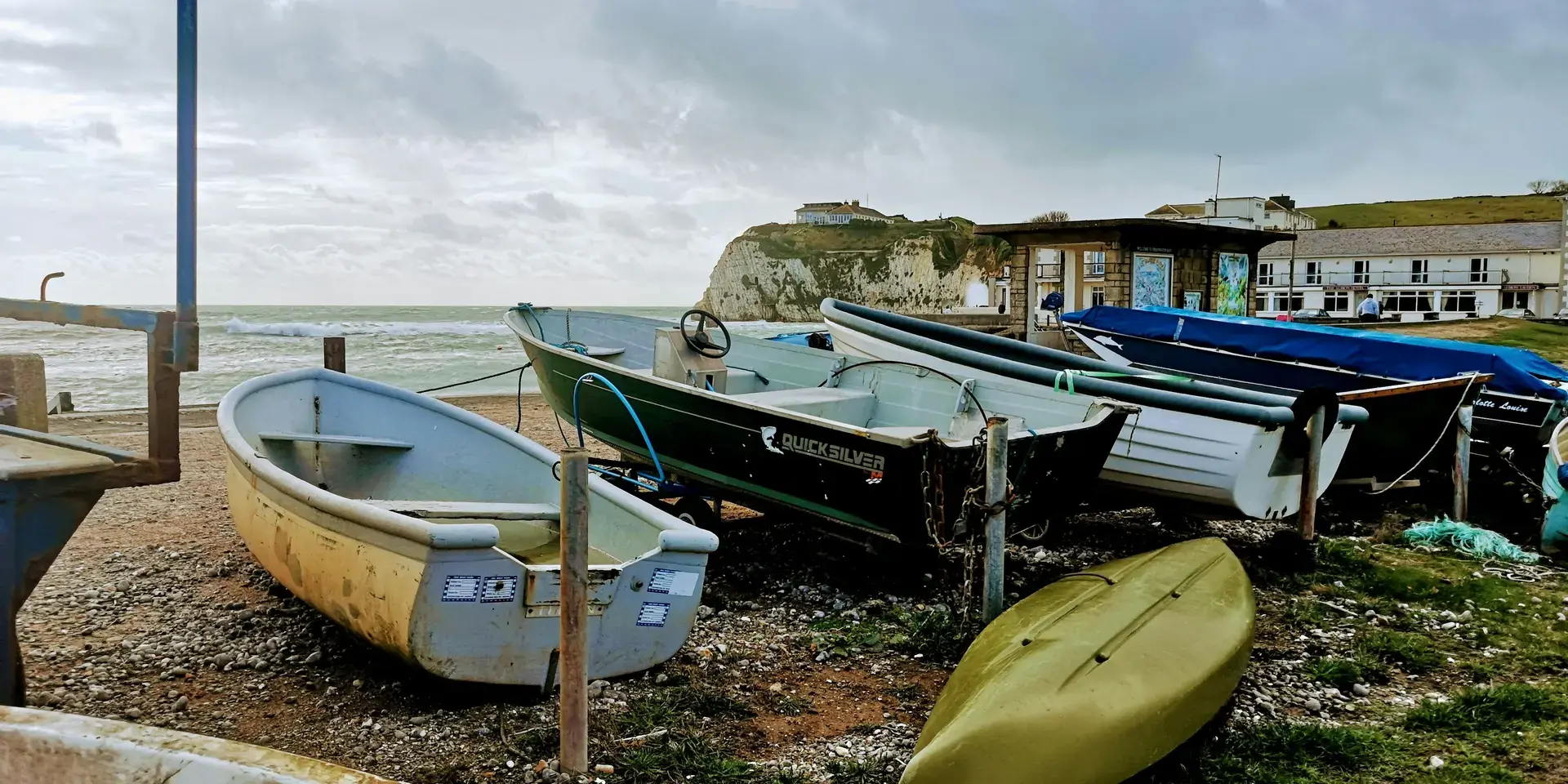 Weathered fishing boats rest on a pebbled shore under a cloudy sky, with cliffs and a calm sea in the background