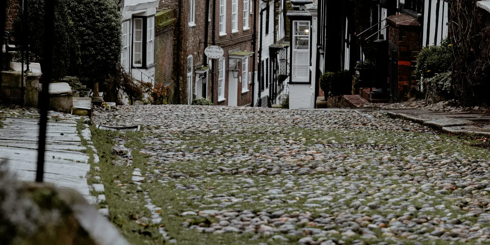 Quaint cobblestone street with mossy stones, historic timber-framed houses, and a gray overcast sky in a hillside village