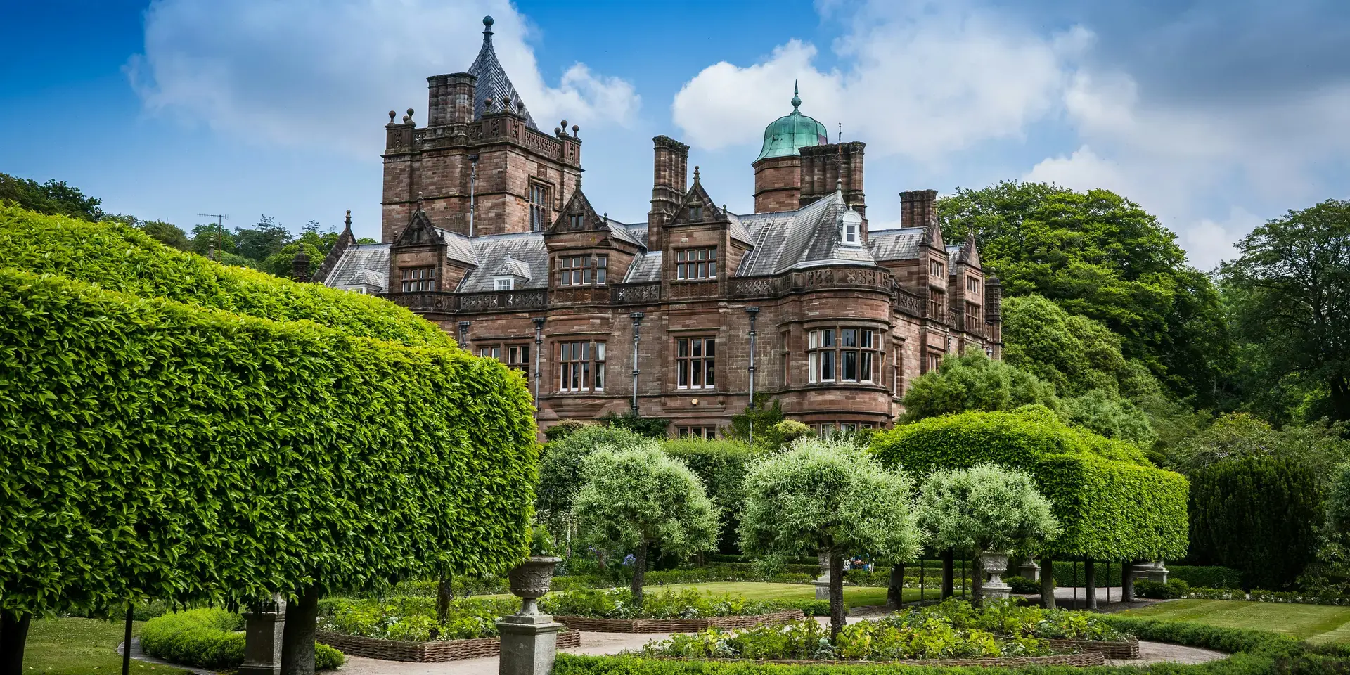 Historic brick manor surrounded by manicured gardens, green hedges, and a bright blue sky with scattered clouds.