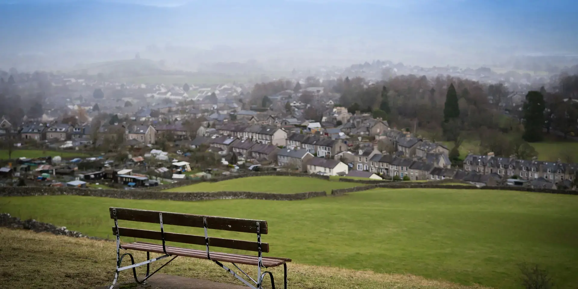 Empty bench overlooking a foggy village surrounded by green fields and distant hills under a pale blue sky