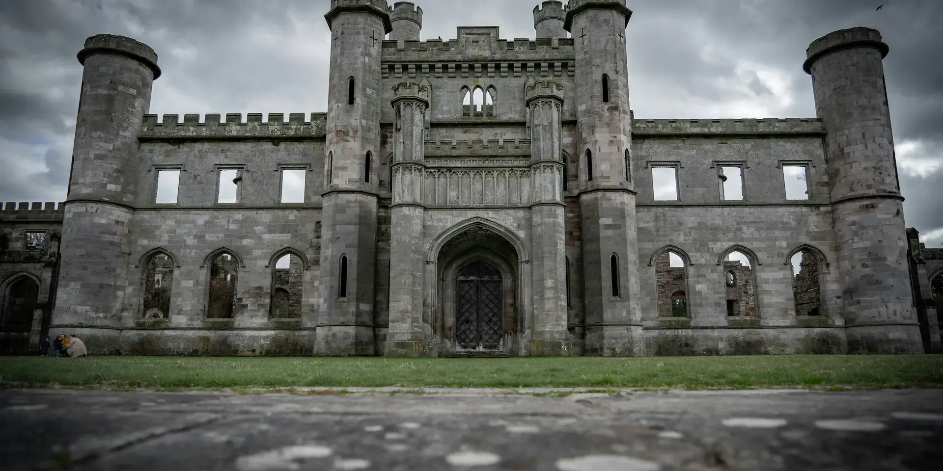 Ruined Gothic-style castle with tall towers and arched windows under a dramatic cloudy sky.