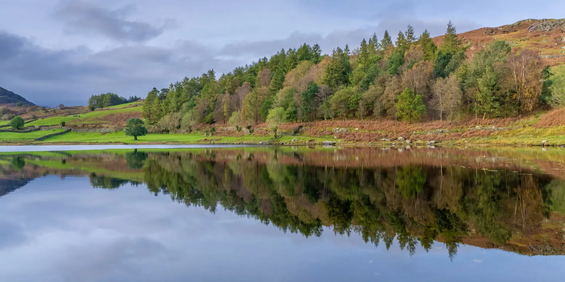 Still lake reflecting trees, green hills, and a cloudy blue sky, surrounded by a peaceful countryside landscape