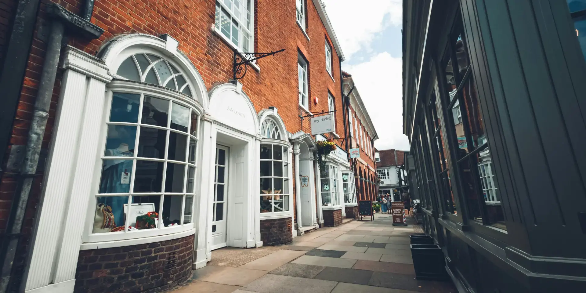 Narrow street lined with red brick buildings and white-framed shop windows, reflecting a quaint and charming town ambiance