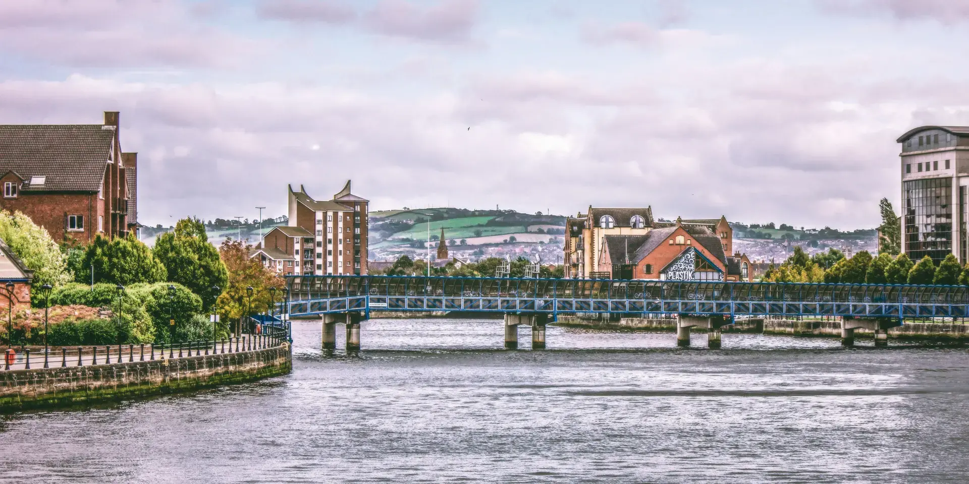 Brick buildings line the riverside with a blue pedestrian bridge spanning the water and rolling green hills in the background