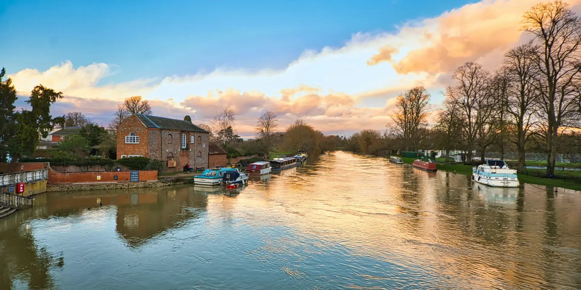 Golden-hour view of a serene river lined with boats, brick houses, and trees,reflecting the vibrant colors of the setting sun