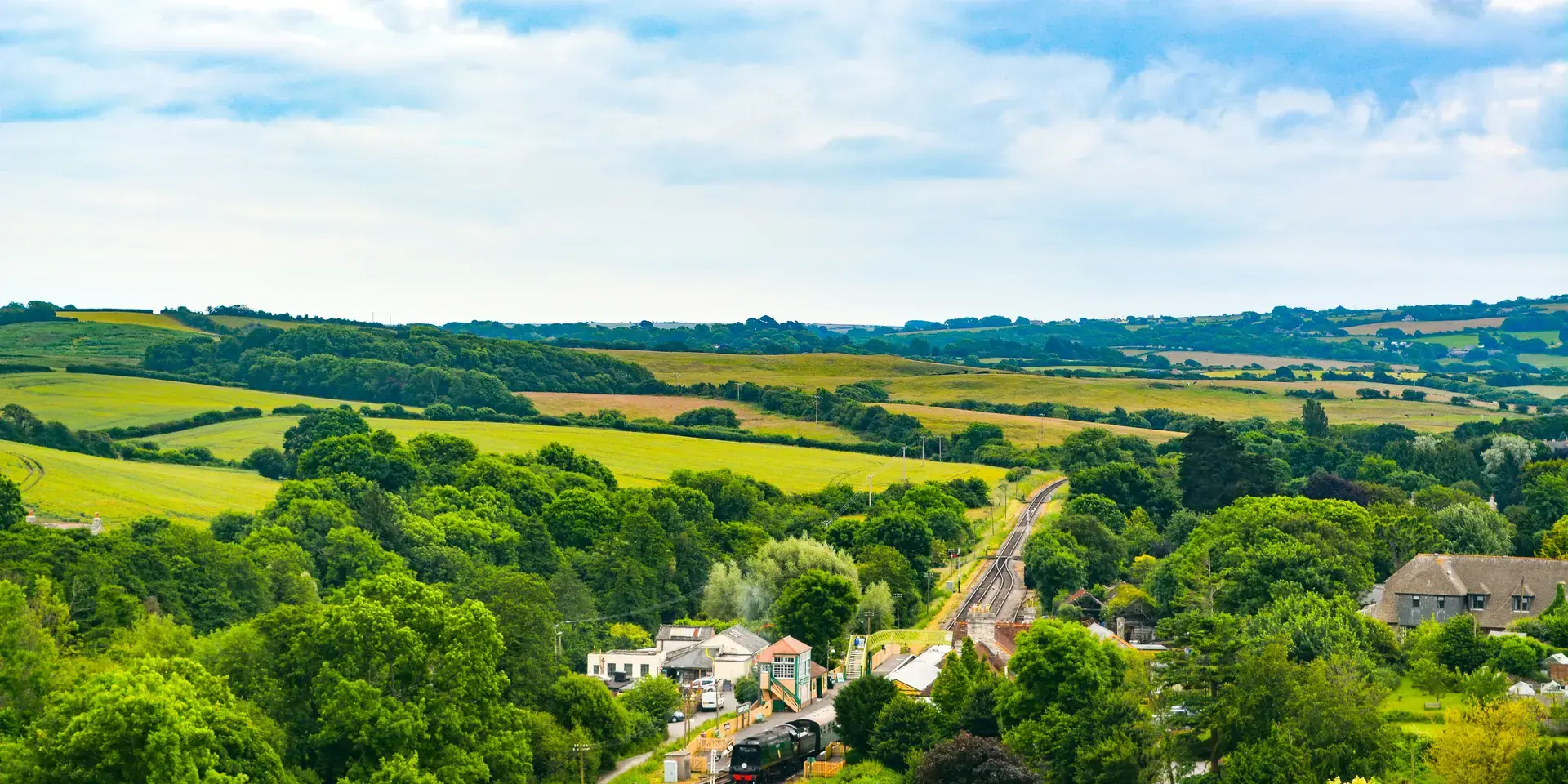 Scenic countryside with green fields, trees, and a small train station nestled between railway tracks under a cloudy sky