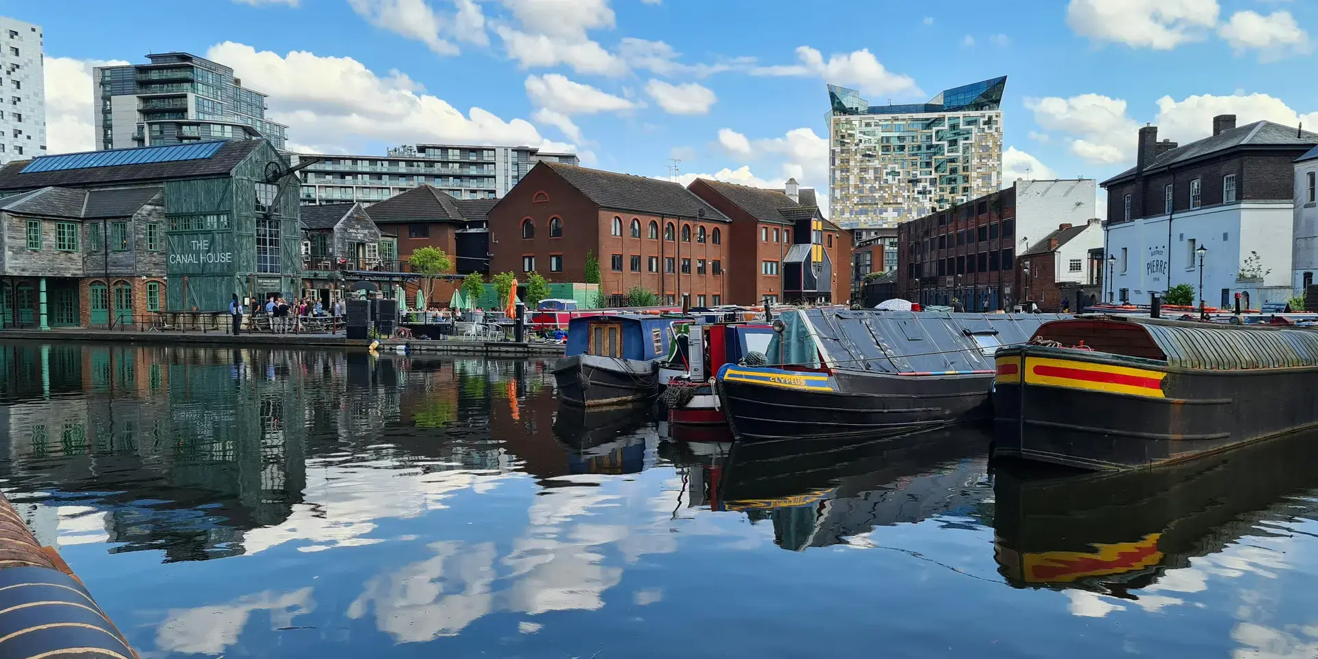 Canal boats docked on calm water, surrounded by historic and modern buildings under a bright blue sky with clouds