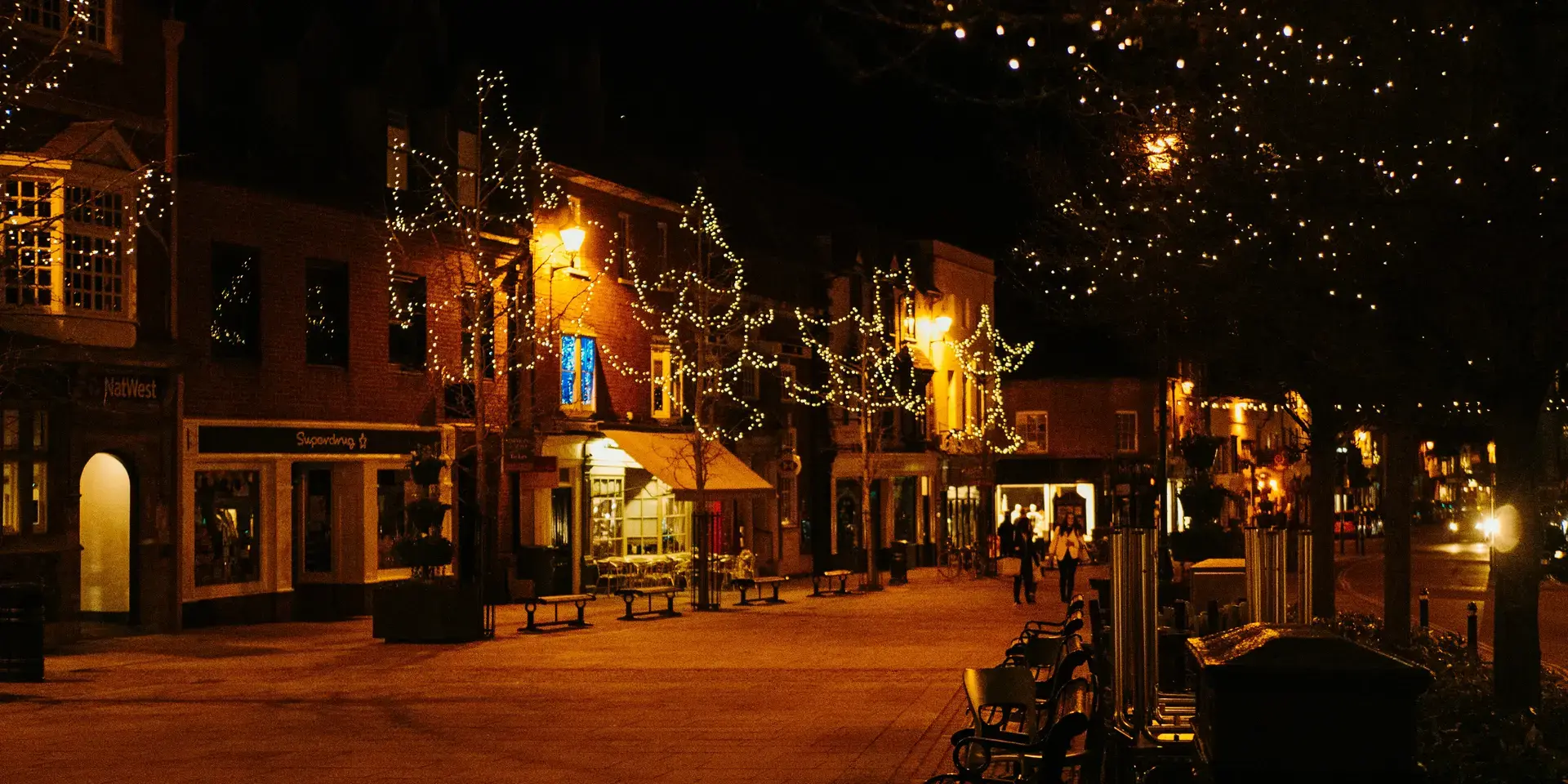 Town square at night with sparkling lights on trees and warm shopfronts, creating a cozy and festive atmosphere