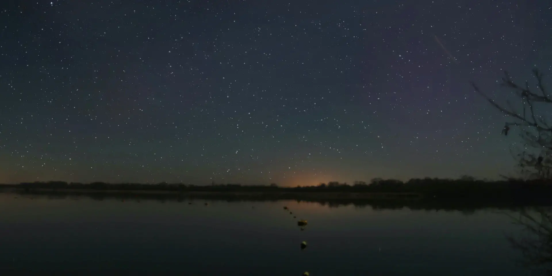 Starry night sky over a calm lake, faint orange glow on the horizon, and a serene atmosphere perfect for stargazing