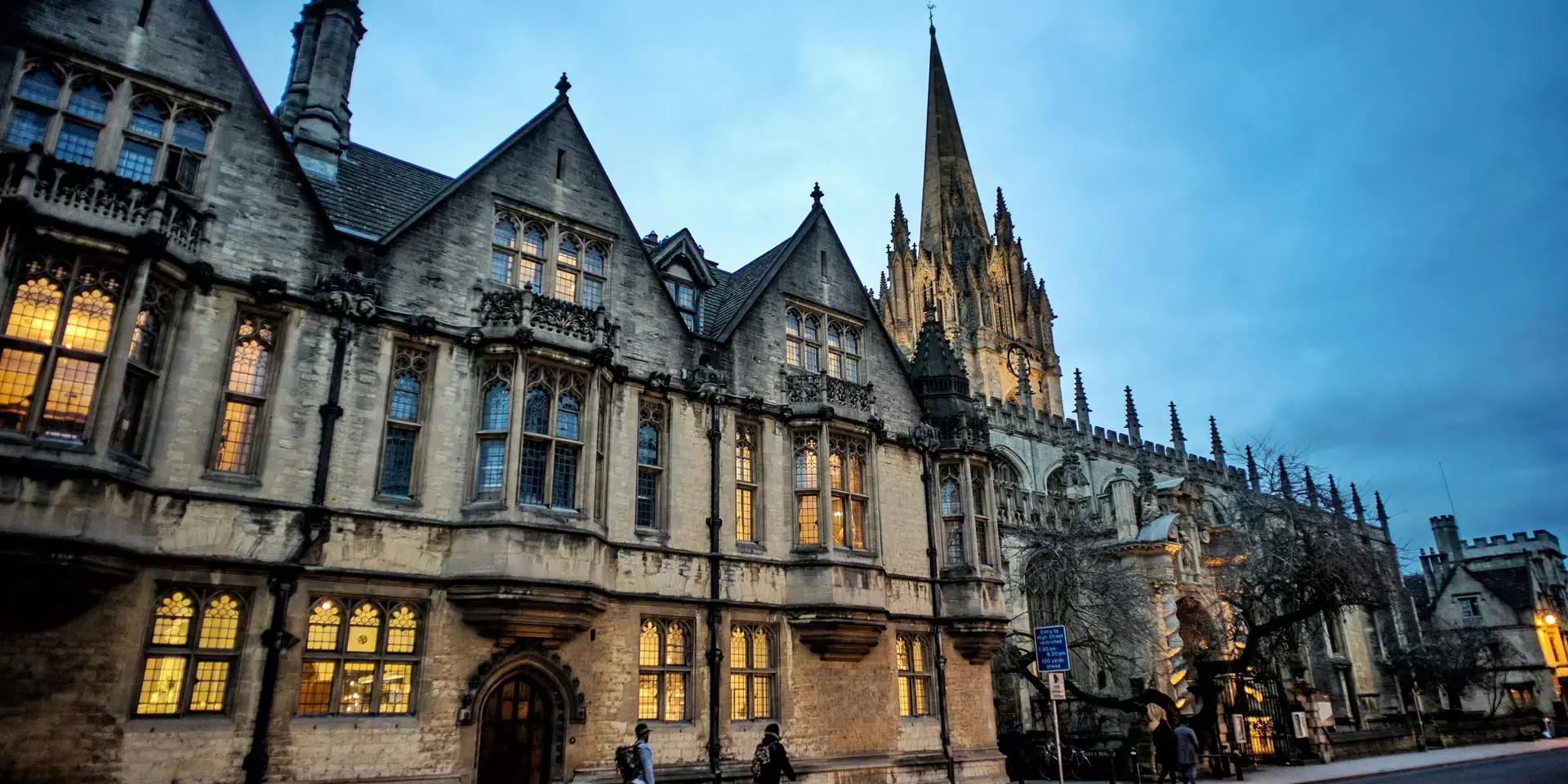 Gothic-style building illuminated at dusk with pointed spires, arched windows, and pedestrians walking along the street
