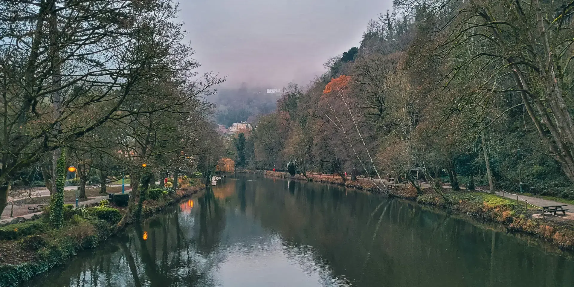 Serene river flanked by bare winter trees and a foggy hillside in the distance, with paths and benches lining the riverbanks
