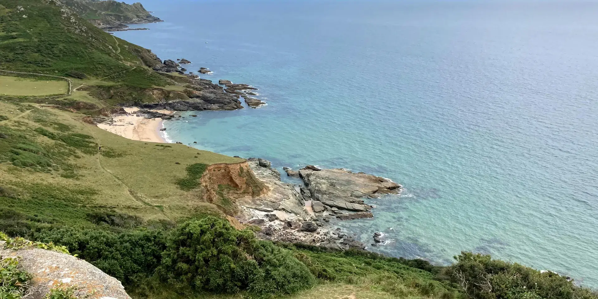Cliffside view of a secluded beach with turquoise waters, rugged coastline, and grassy hills under a sunny blue sky