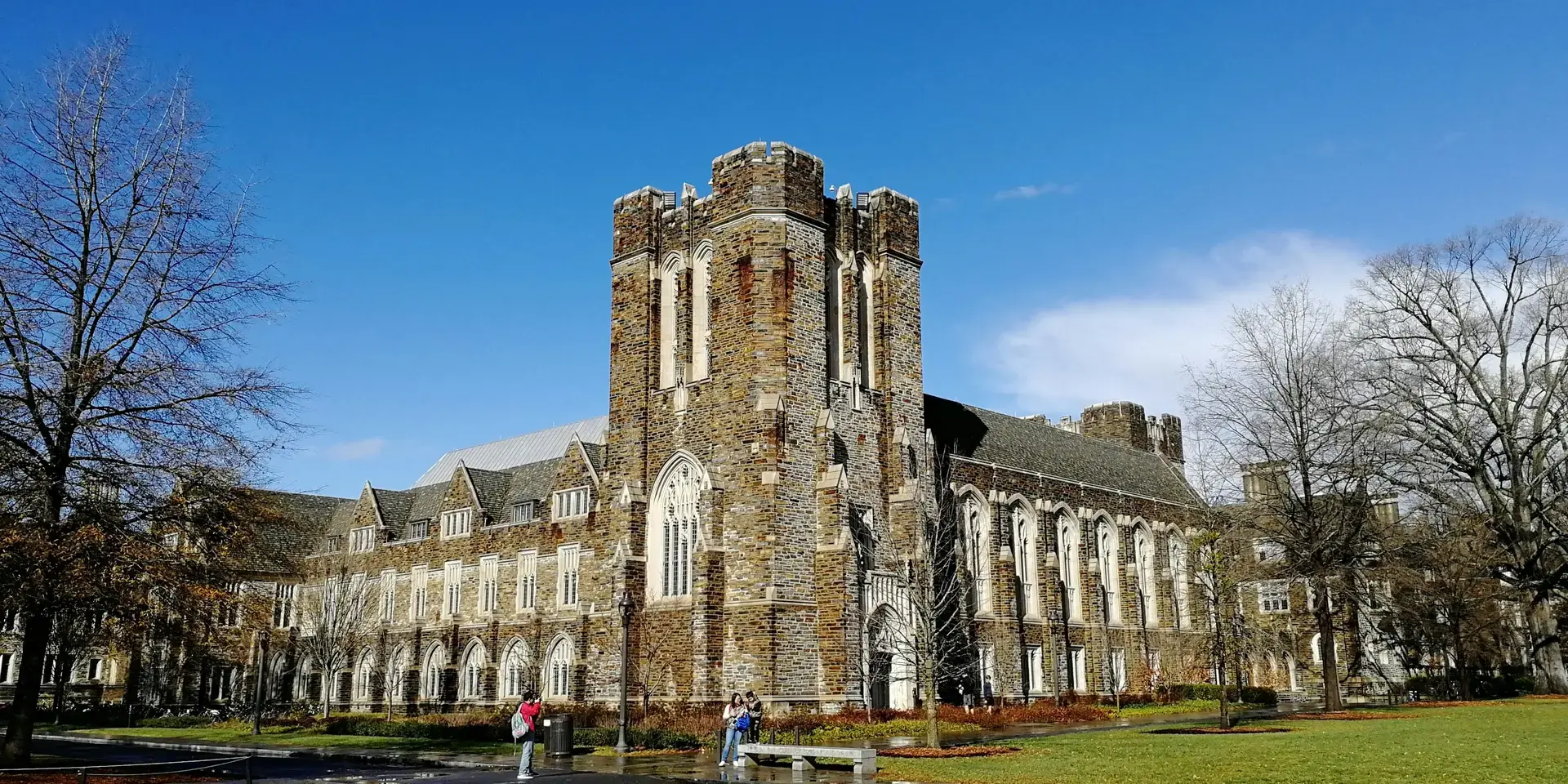 Gothic-style stone building with arched windows, central tower, green lawn, and blue sky.