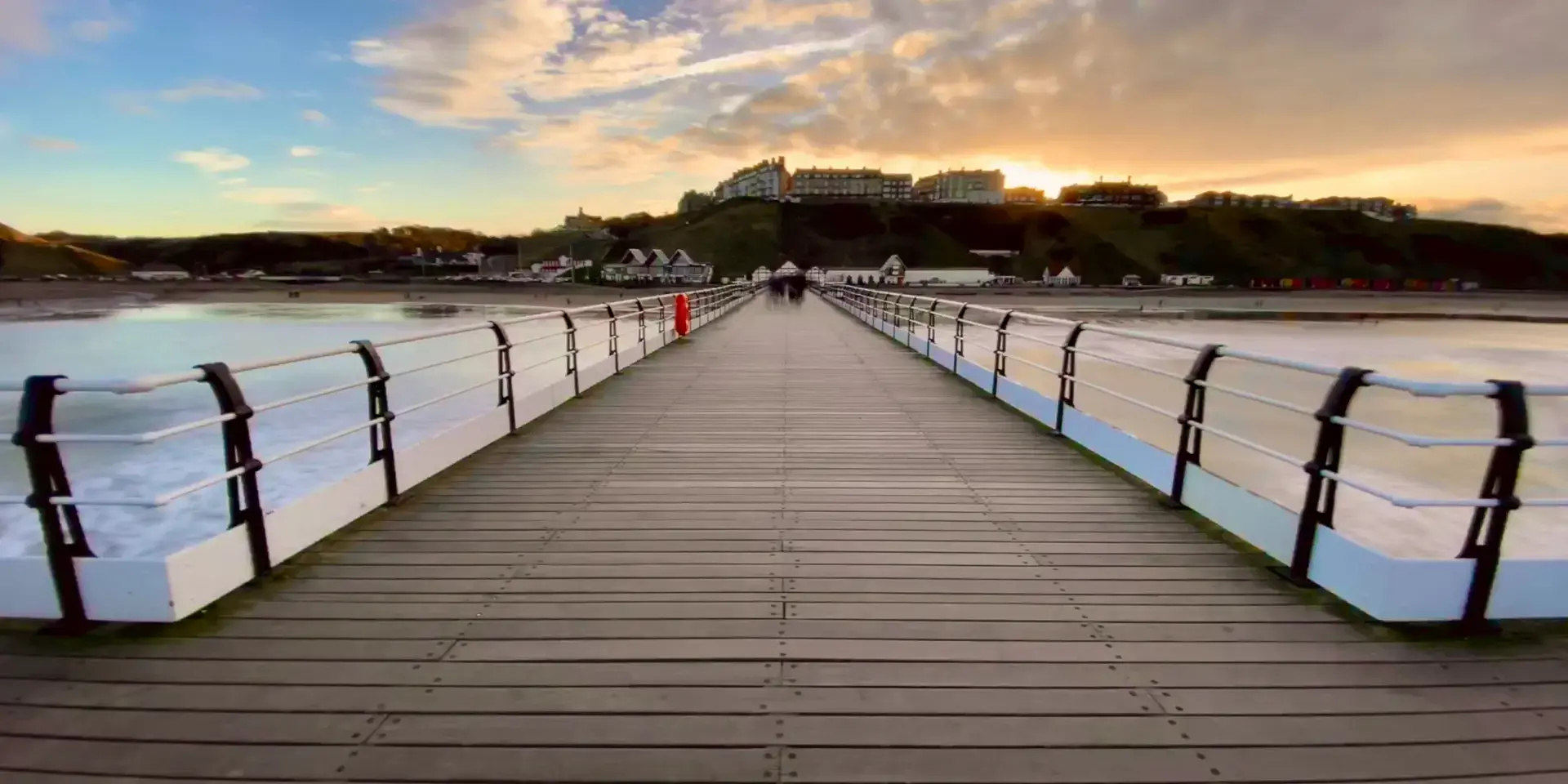 Wooden pier extending towards the horizon, flanked by calm waters and hills, with a glowing sunset illuminating the sky