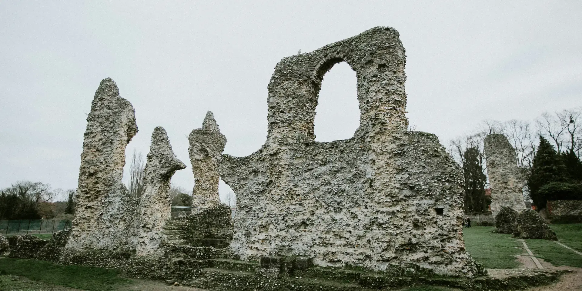 Ancient stone ruins with weathered textures stand on a grassy field under an overcast sky, surrounded by sparse winter trees