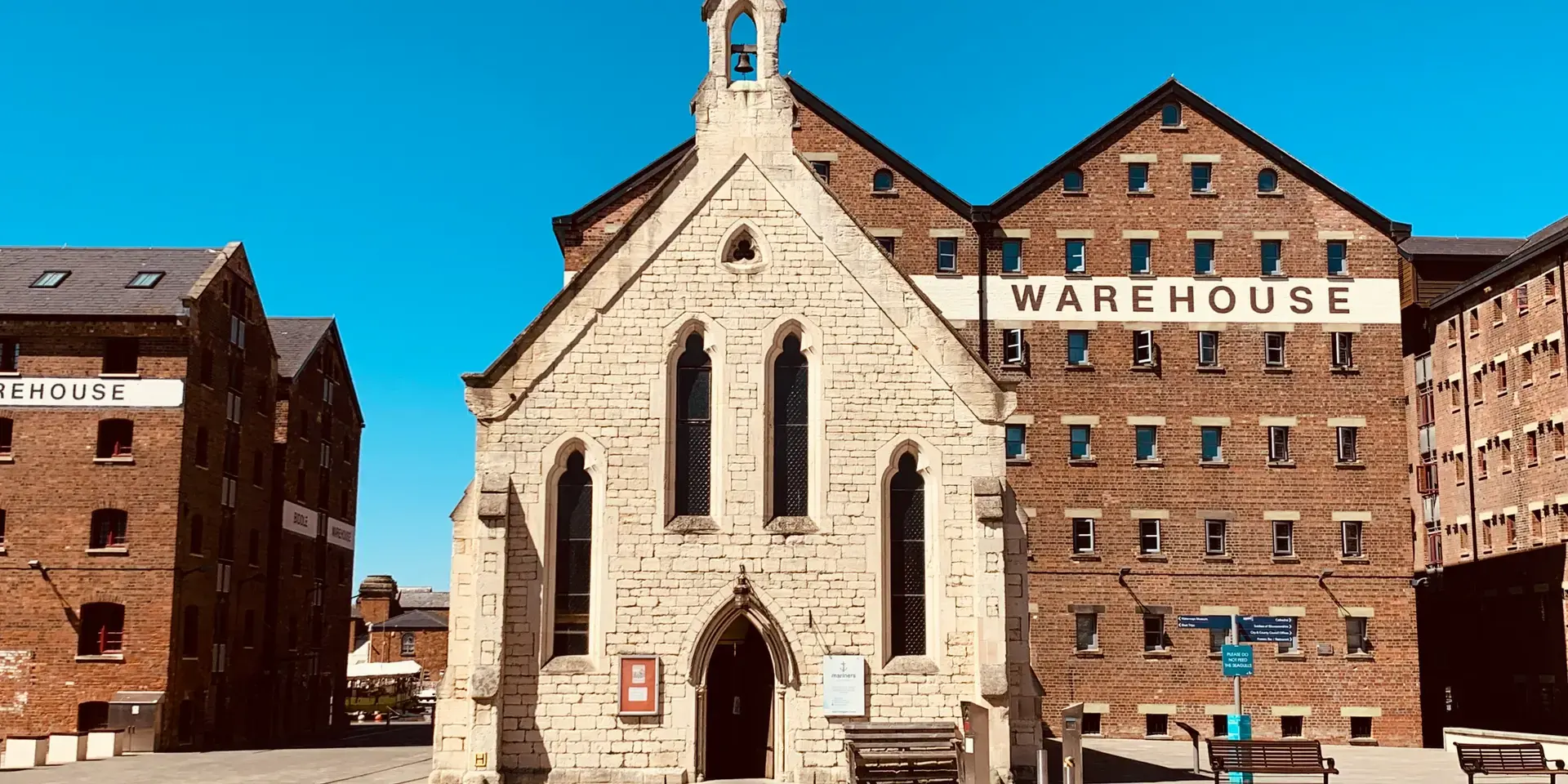 Historic stone chapel surrounded by red-brick warehouses under a bright blue sky, blending tradition with industrial heritage