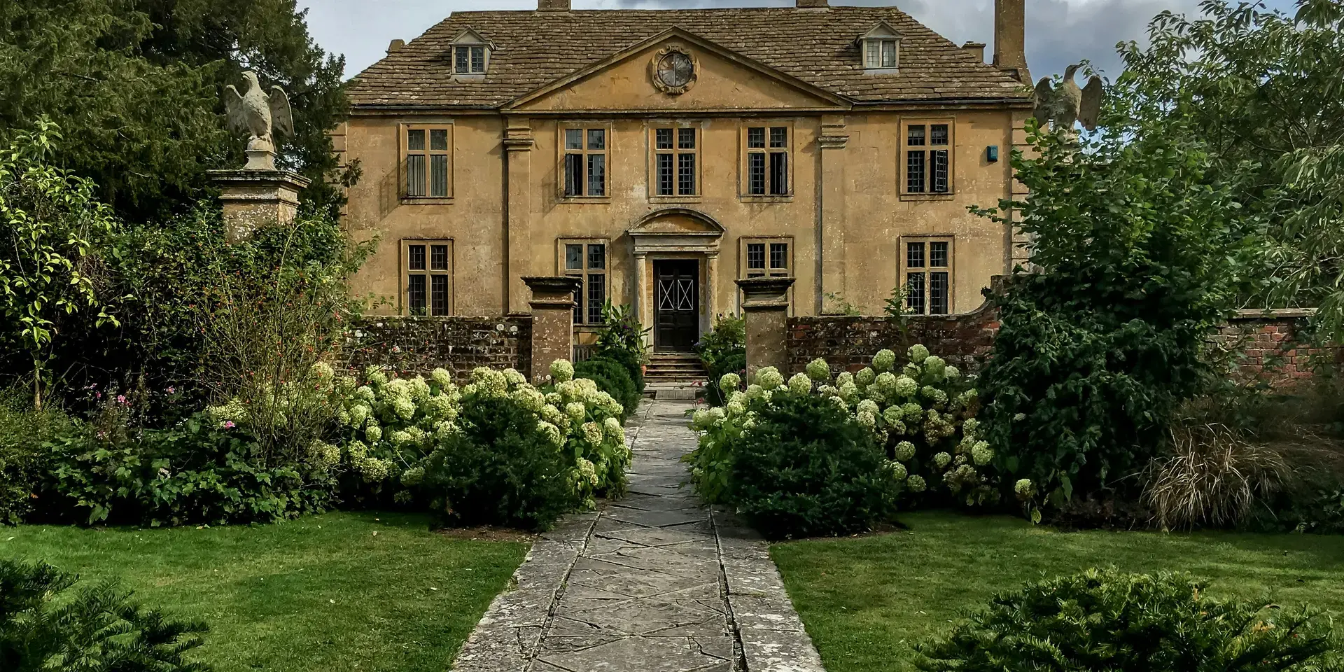 Elegant historic manor house with a stone pathway, surrounded by lush gardens and hedges under a cloudy sky