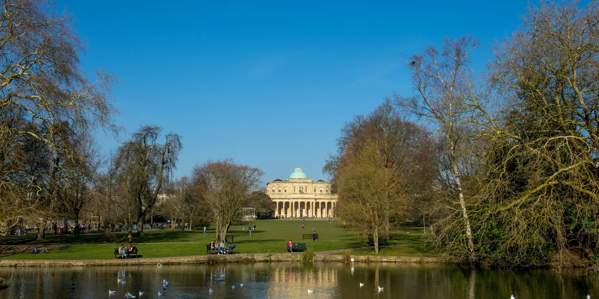 Reflections in a calm park lake surrounded by leafless trees, with a neoclassical building under a vivid blue sky