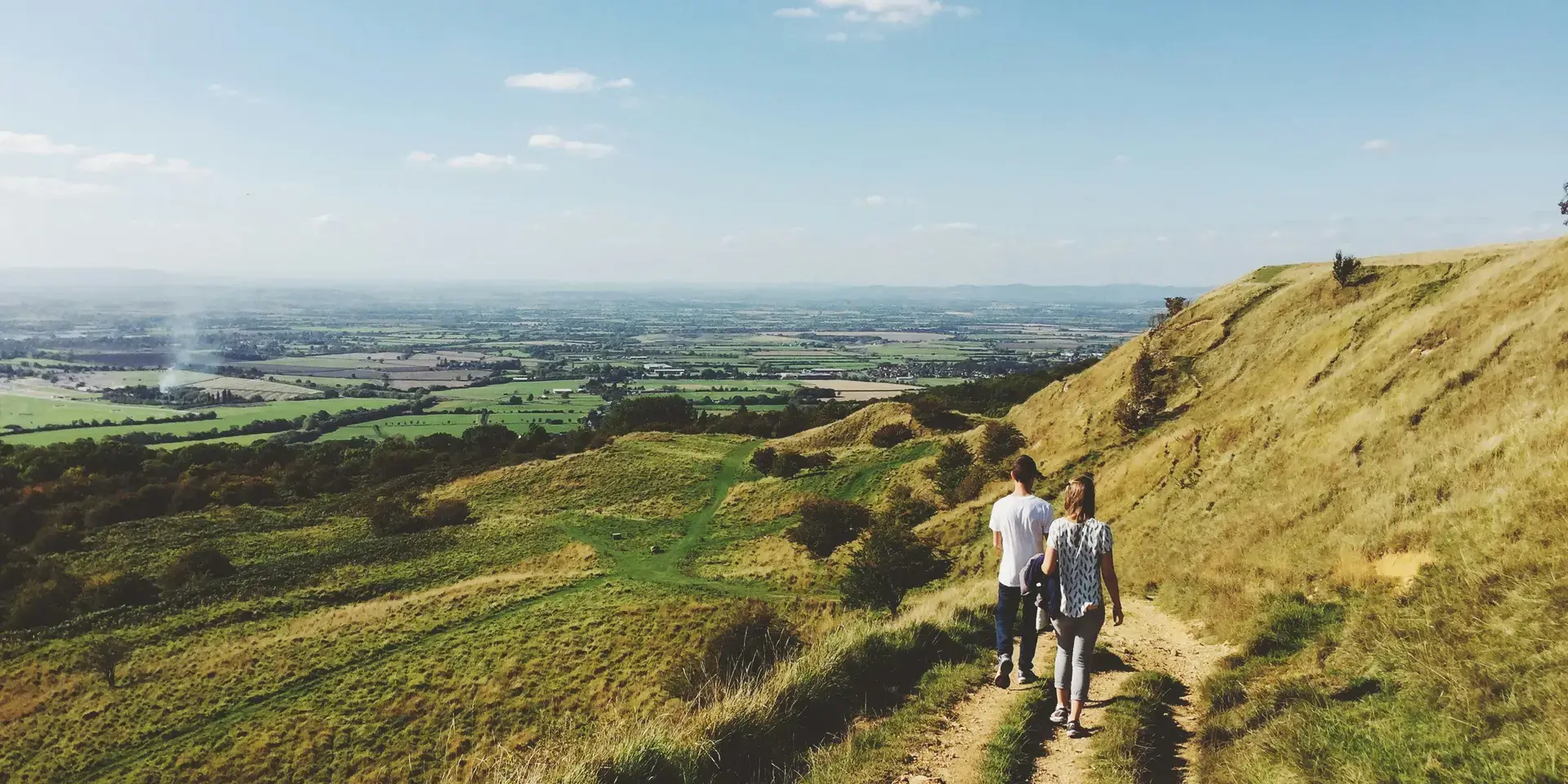 Two hikers walk a hillside trail overlooking green fields and a blue sky, surrounded by breathtaking natural scenery