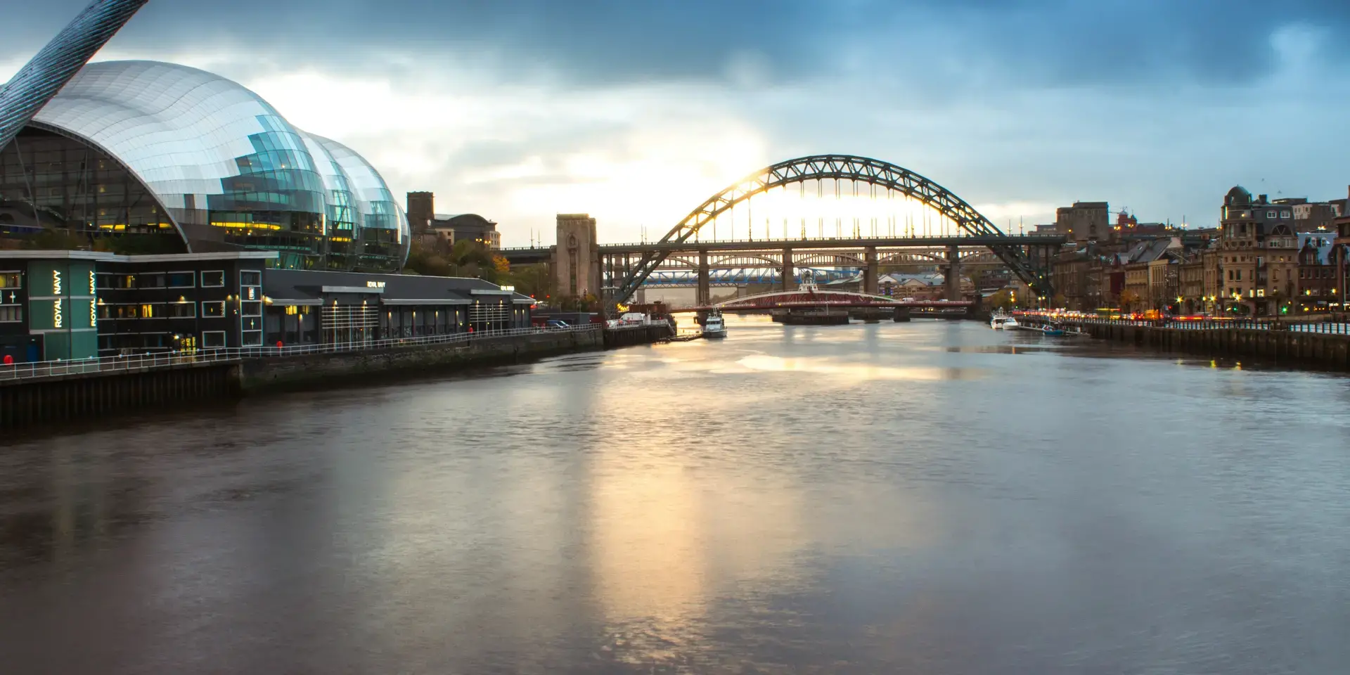 Evening view of a modern riverside with a glass dome building, iconic arched bridge, and reflections on the calm water