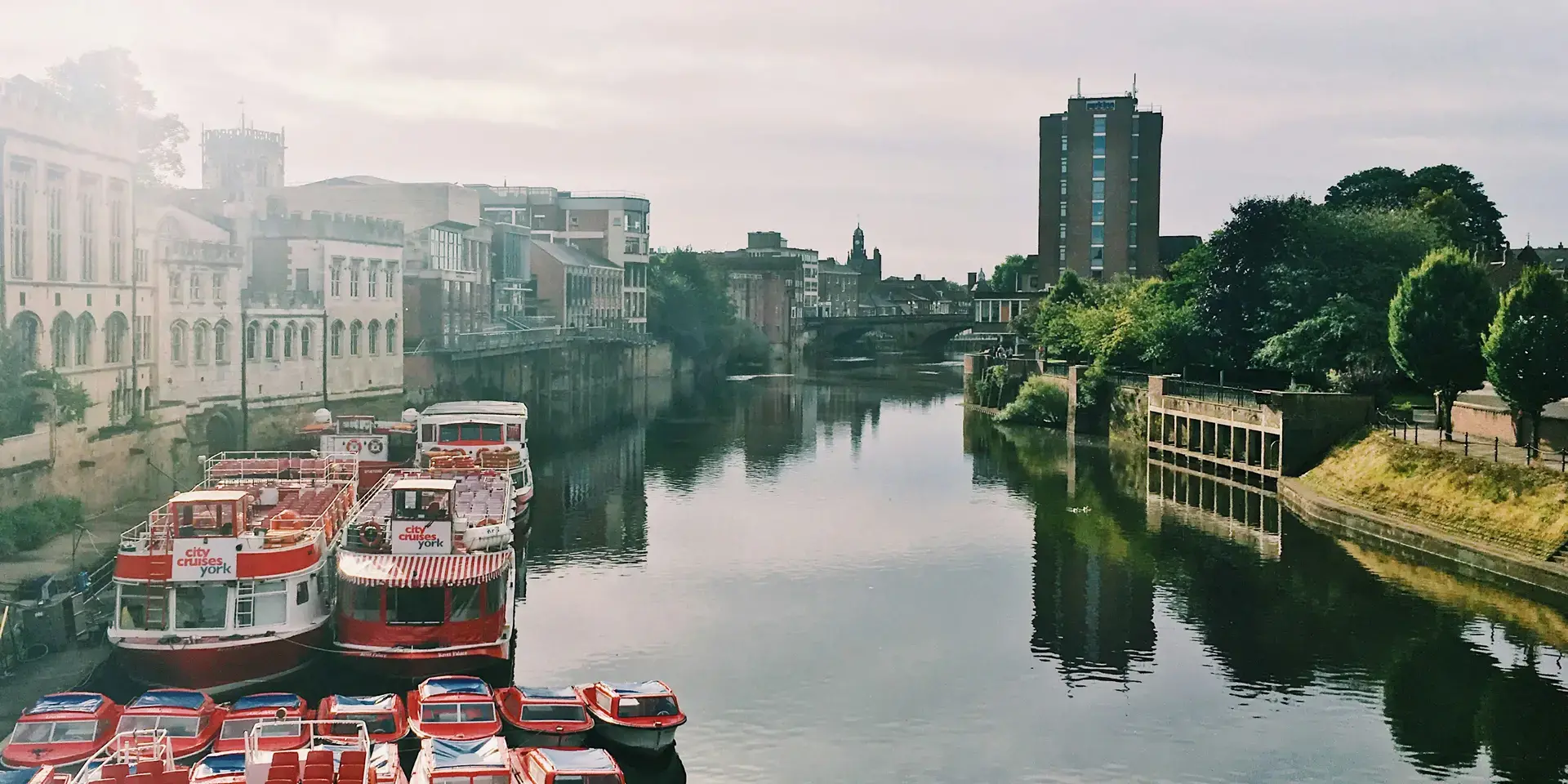 Colorful boats moored on a tranquil river with historic buildings and modern structures under a soft cloudy sky