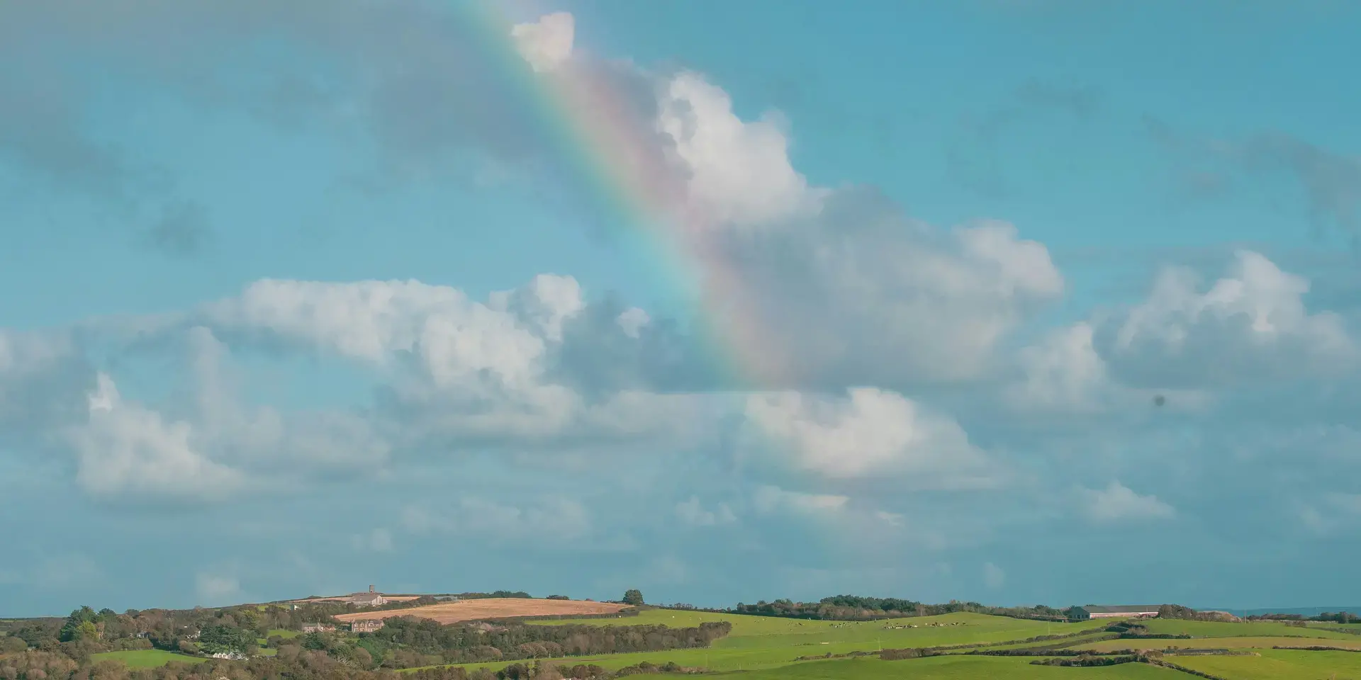 Vivid rainbow arches over lush green fields and a serene estuary dotted with small boats under a bright blue sky