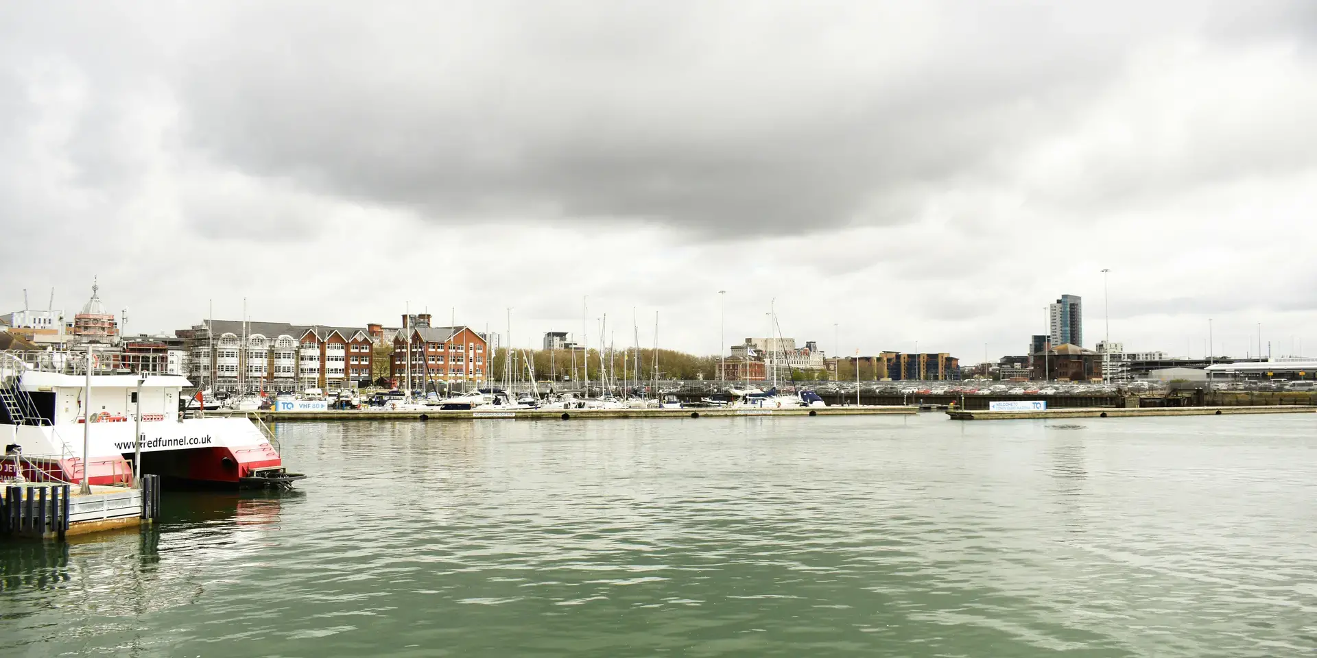 Marina with moored boats under cloudy skies, surrounded by modern and traditional buildings reflecting on calm green water