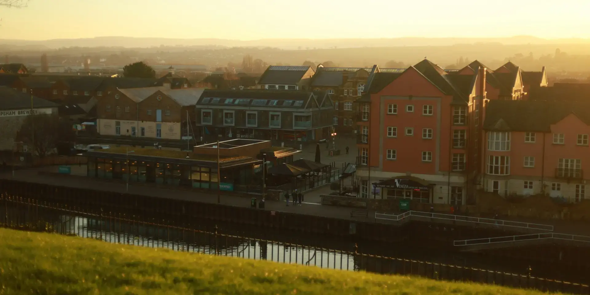 Urban riverside area with modern buildings, soft evening sunlight, and a grassy foreground overlooking the scene
