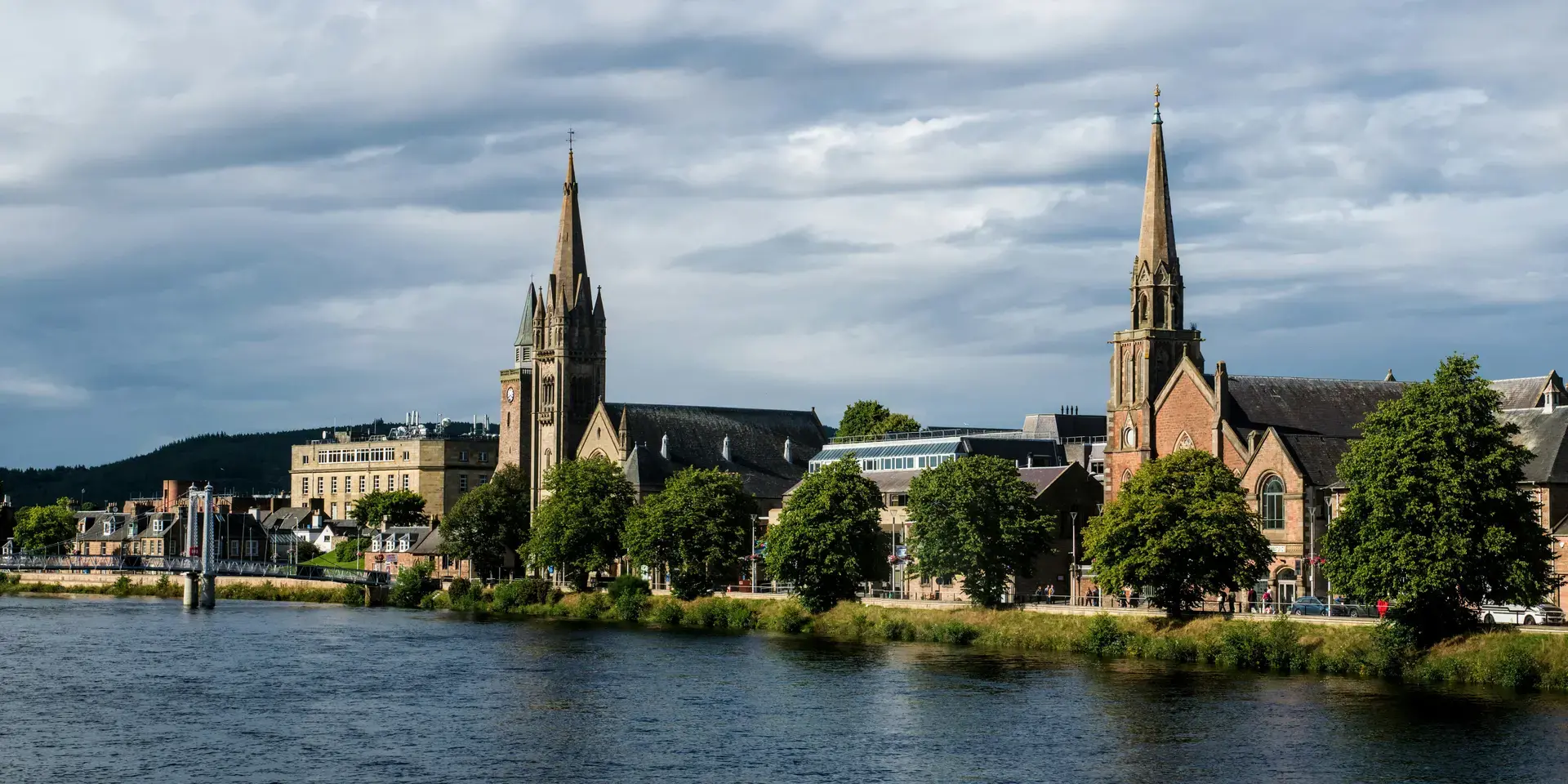 Historic riverside buildings with tall spires under cloudy skies, framed by greenery and a scenic waterfront