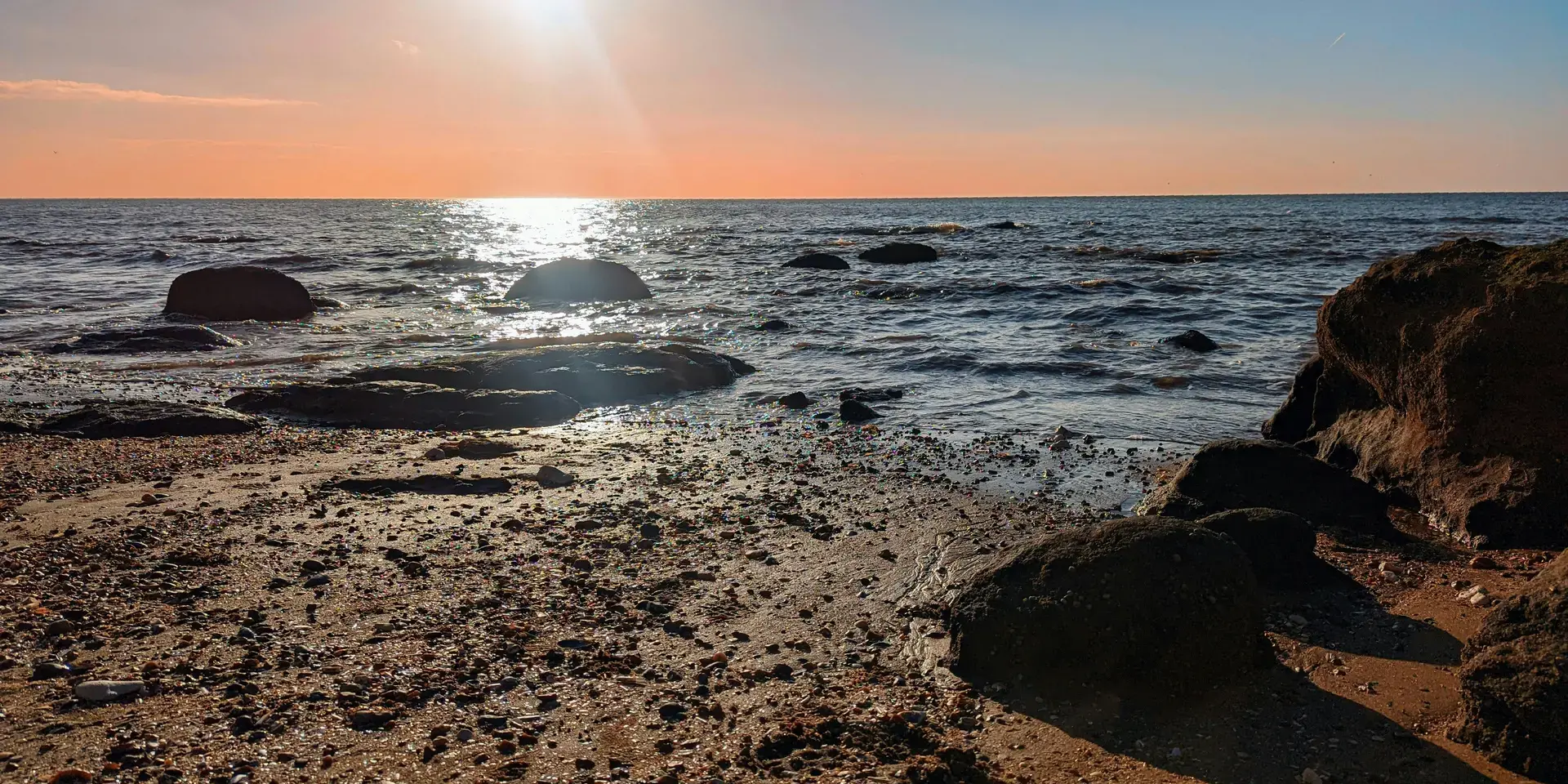 Rocky shoreline at sunset, with golden light reflecting off the sea and scattered pebbles on the sandy beach