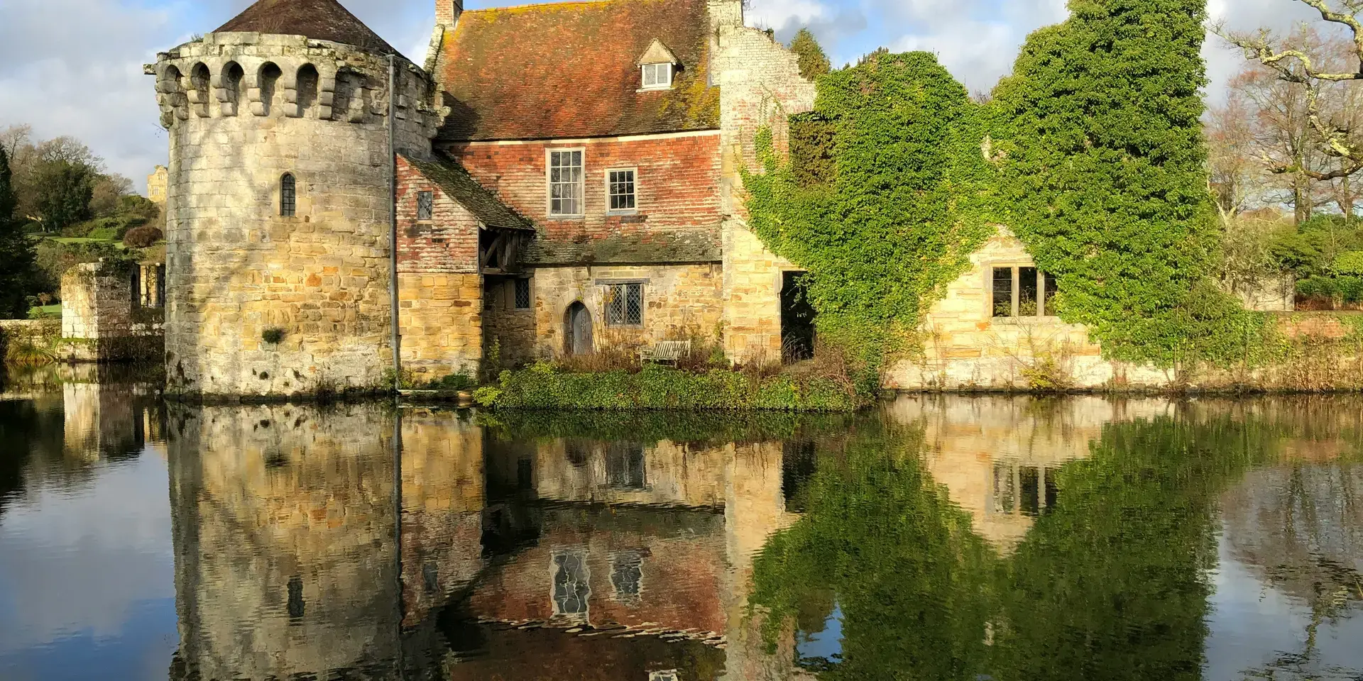 Historic stone castle with ivy-covered walls reflecting in a calm moat, under a partly cloudy sky